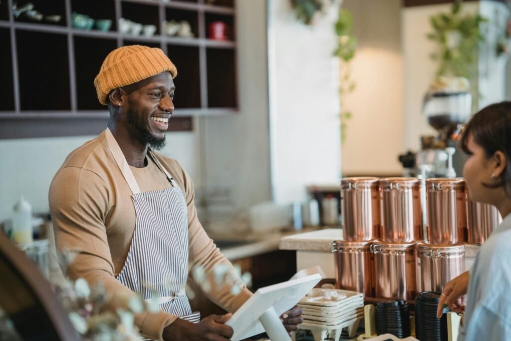 Smiling barista wearing an apron and beanie engaging with a customer at a café counter.