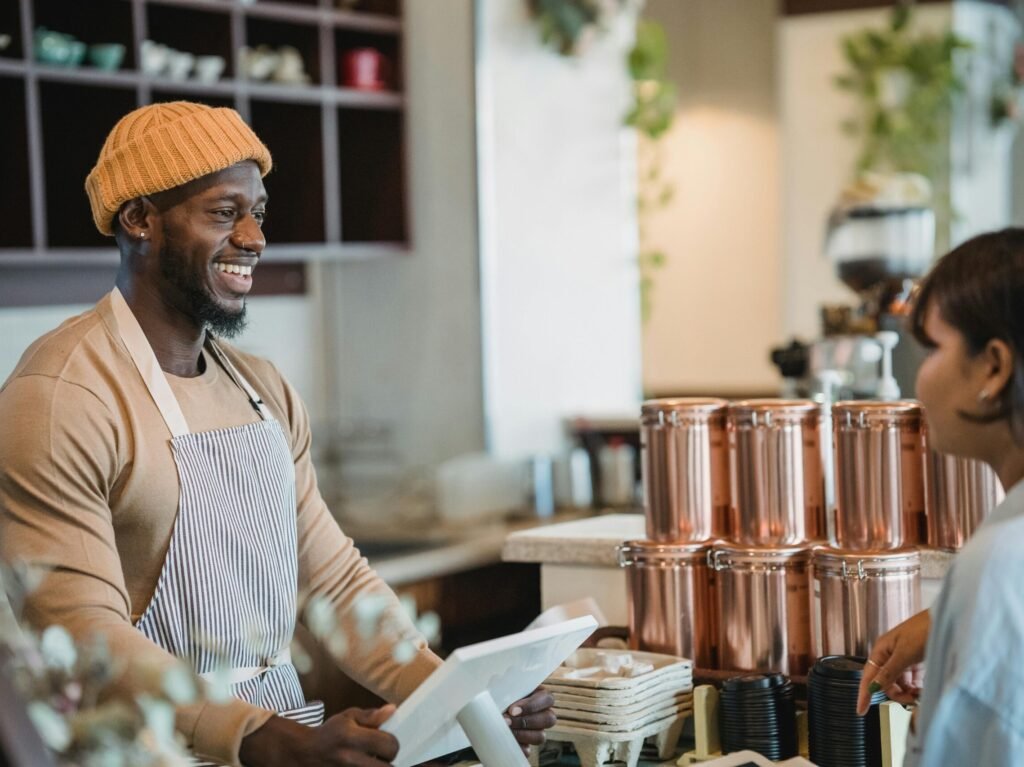 peolple of Color: Smiling barista wearing an apron and beanie engaging with a customer at a café counter.wearing a beige shirt