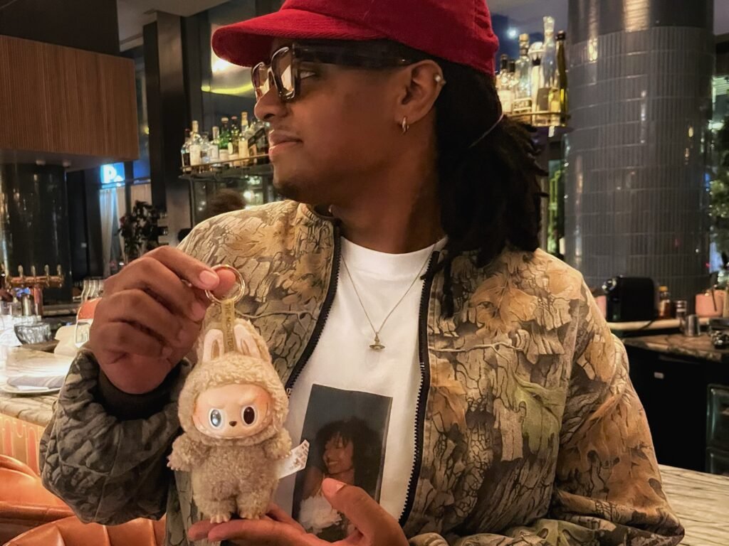 young person of colour wearing large glasses and a red cap with Labubu doll